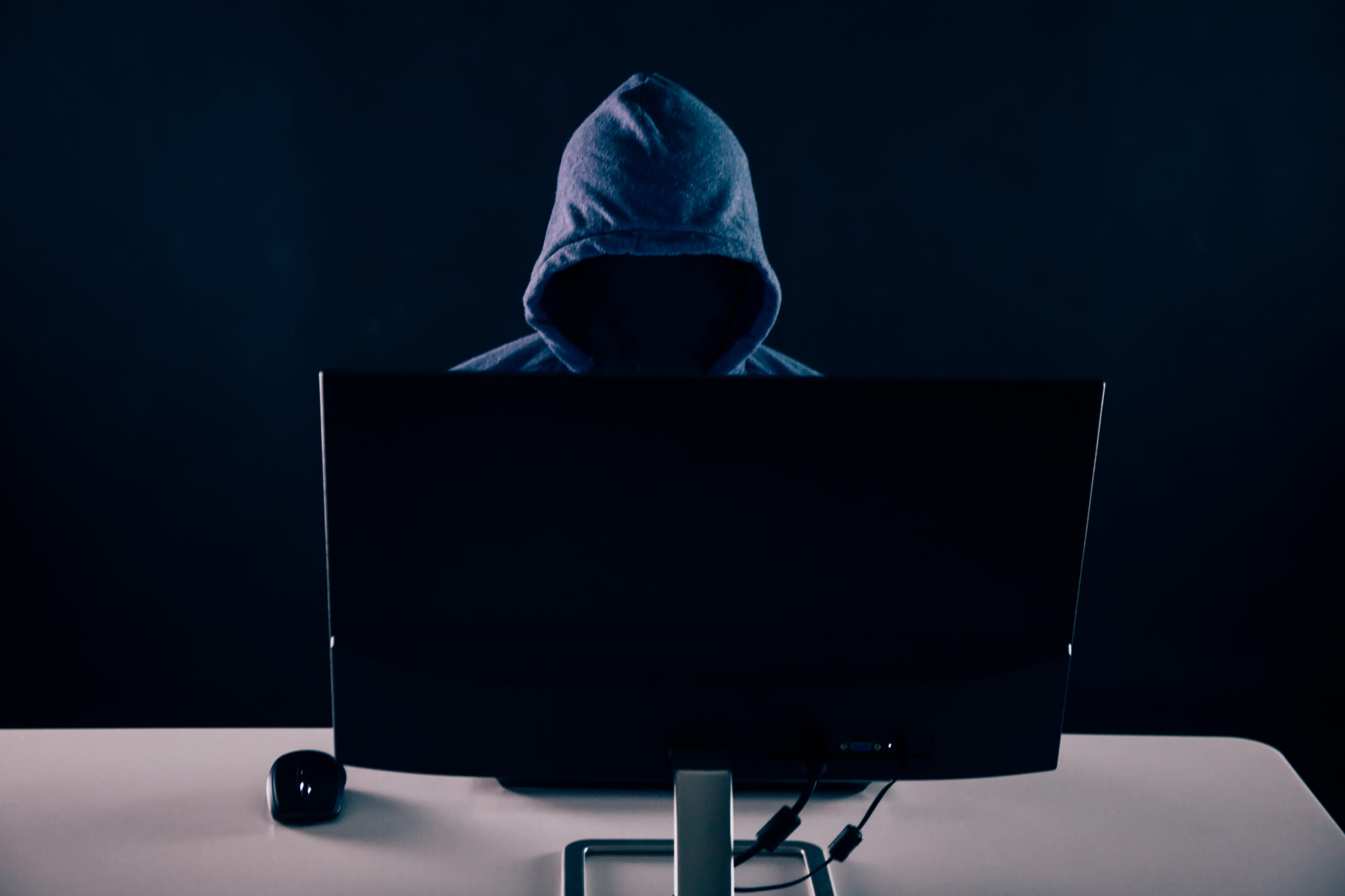 Anonymous and faceless hacker under hoodie using computer isolated over dark background - illegal online internet criminal concept